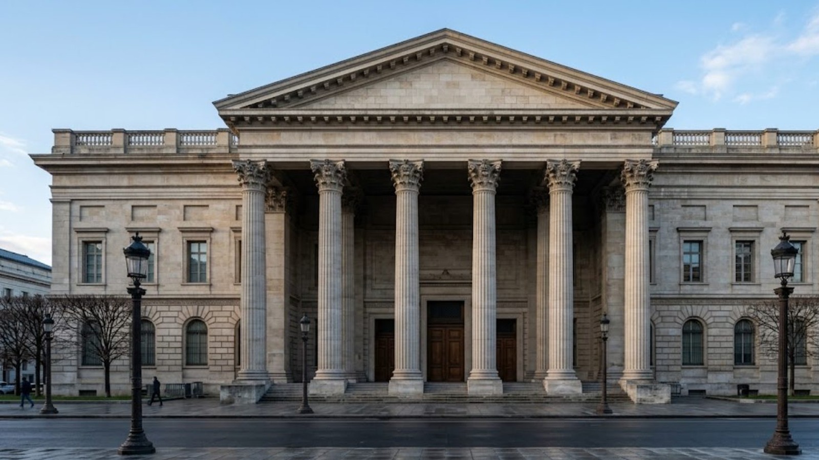 A neoclassical government building with a columned portico at first light, an empty plaza and wet road in front of it, no people