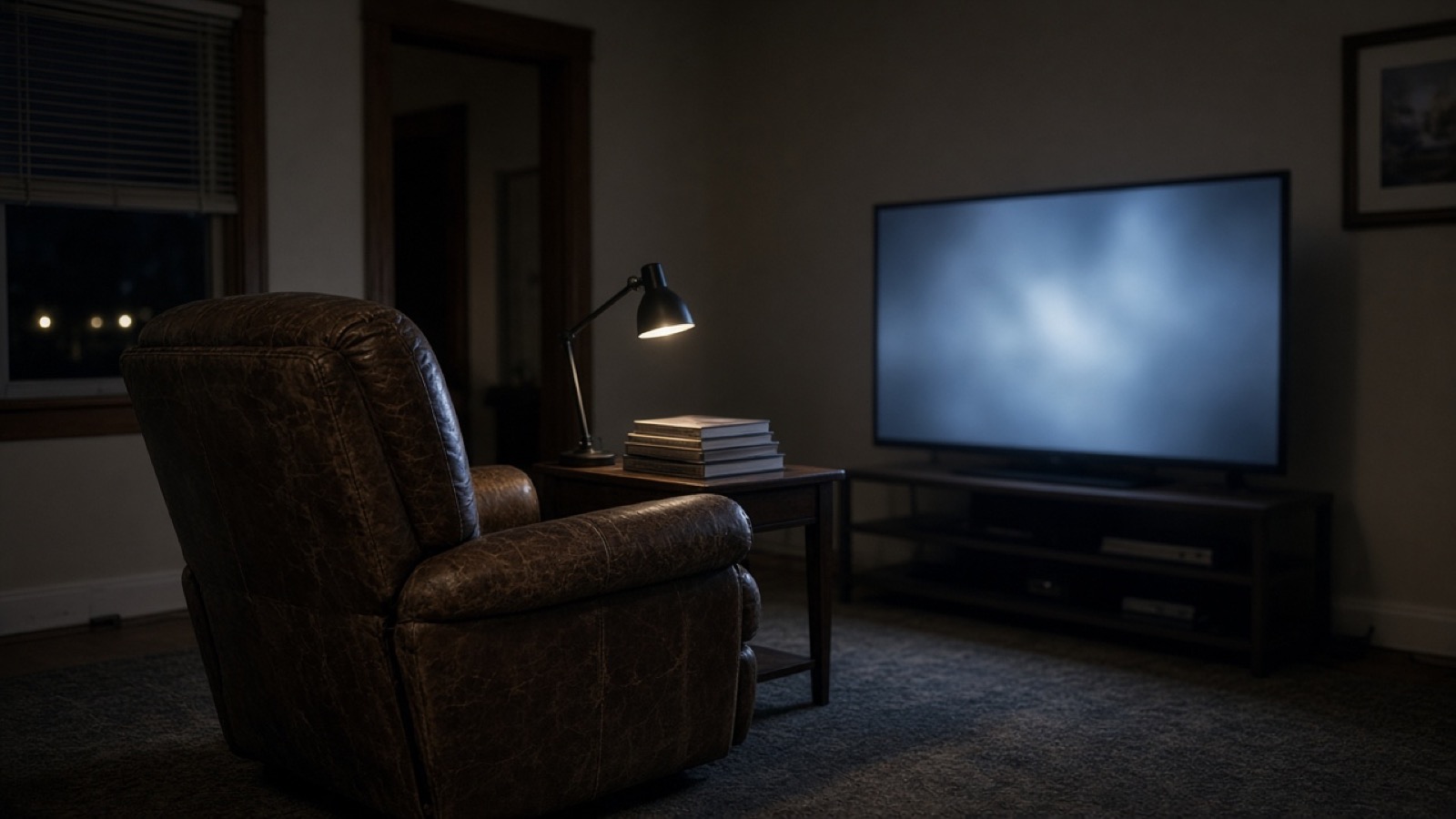 A worn brown leather recliner faces a large flat-screen television glowing pale blue in a dim living room at night, a small desk lamp lighting a stack of books on a side table beside it.