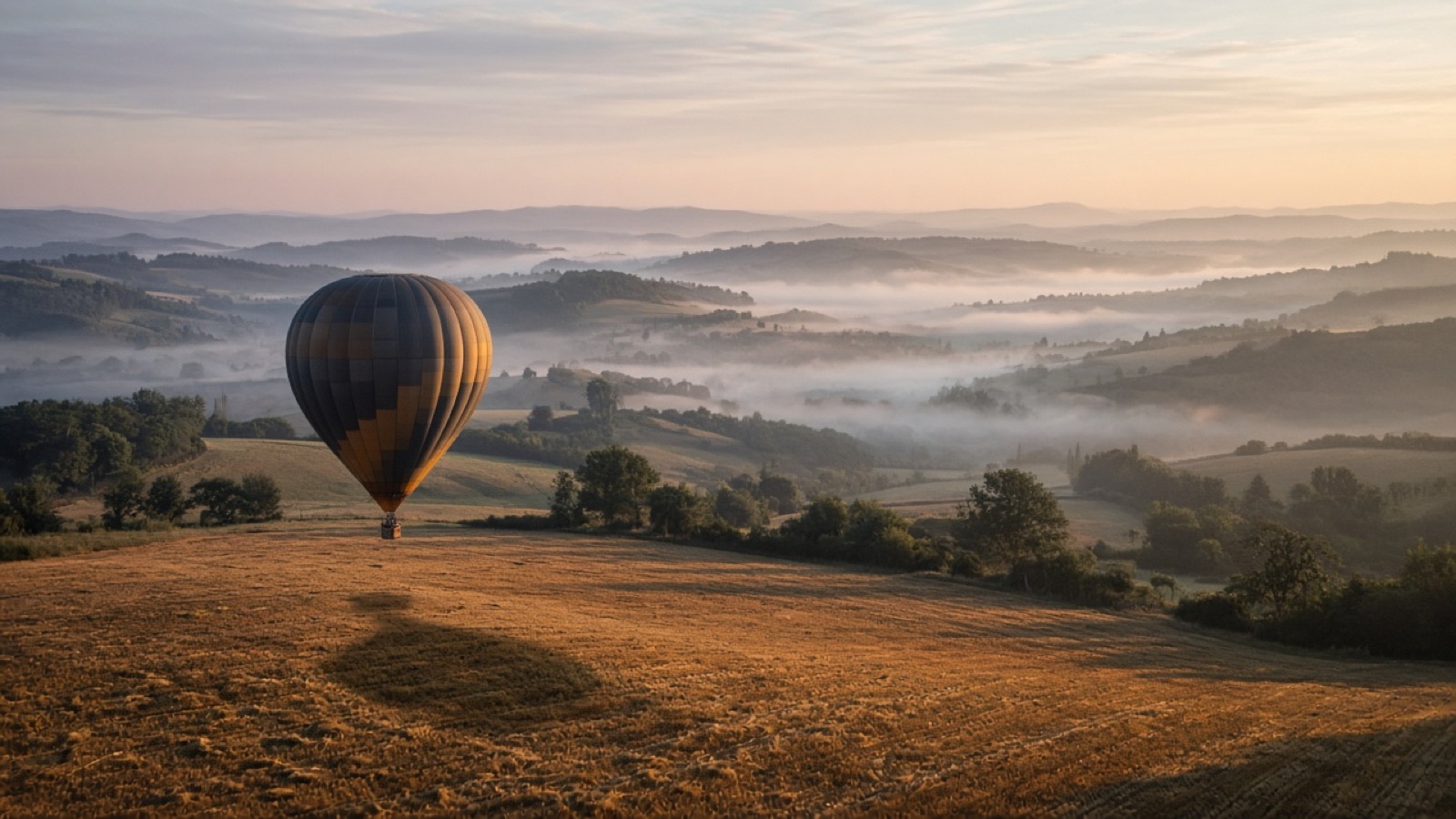 A single dark-and-amber hot-air balloon floats low over a harvested golden field at dawn, its long shadow on the stubble, with misty rolling hills and wooded valleys fading under a pale pink sky.