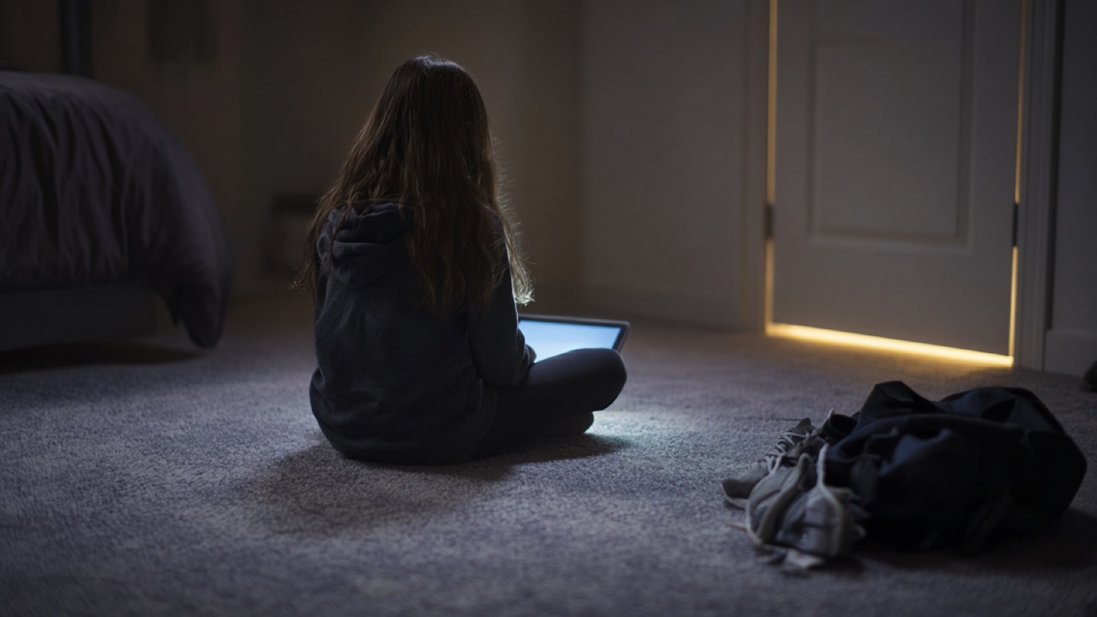 A girl with long hair, seen from behind, sits cross-legged on a bedroom carpet in the dark, lit by the cool glow of a tablet, while warm hallway light leaks around a closed door near a dropped bag and sneakers.