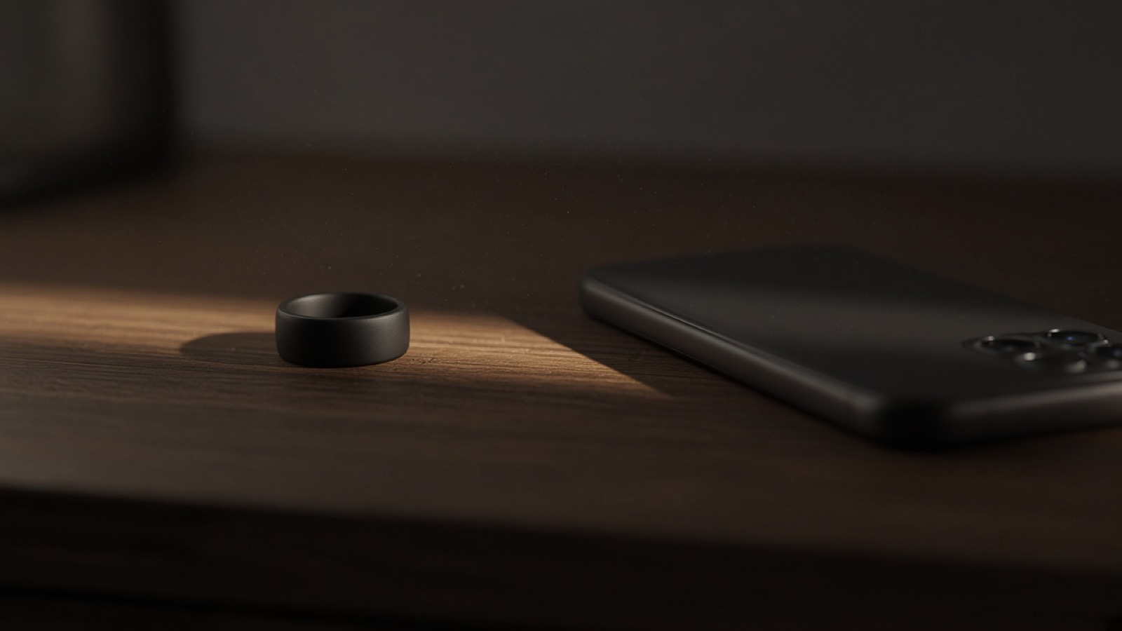 A matte black ring rests on a dark wooden nightstand in a thin band of early morning light, beside a black smartphone lying face down, its camera lenses facing up, in an otherwise dim, quiet room.