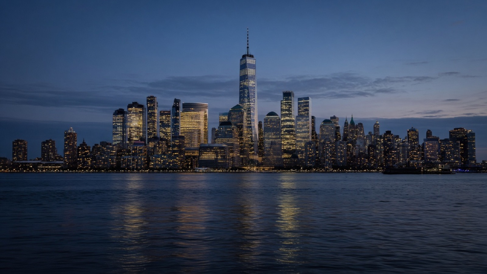 The lit towers of Lower Manhattan's financial district rise across calm dark harbor water at blue hour, the tallest spire at center, with thousands of glowing office windows reflected on the surface.