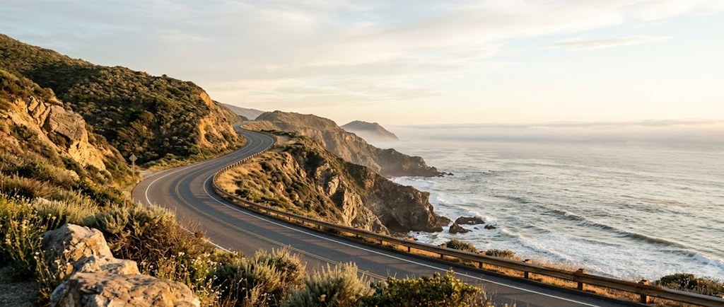A two-lane coastal road curves along brush-covered bluffs above rocky surf at golden hour, a low fog bank resting on the ocean horizon.