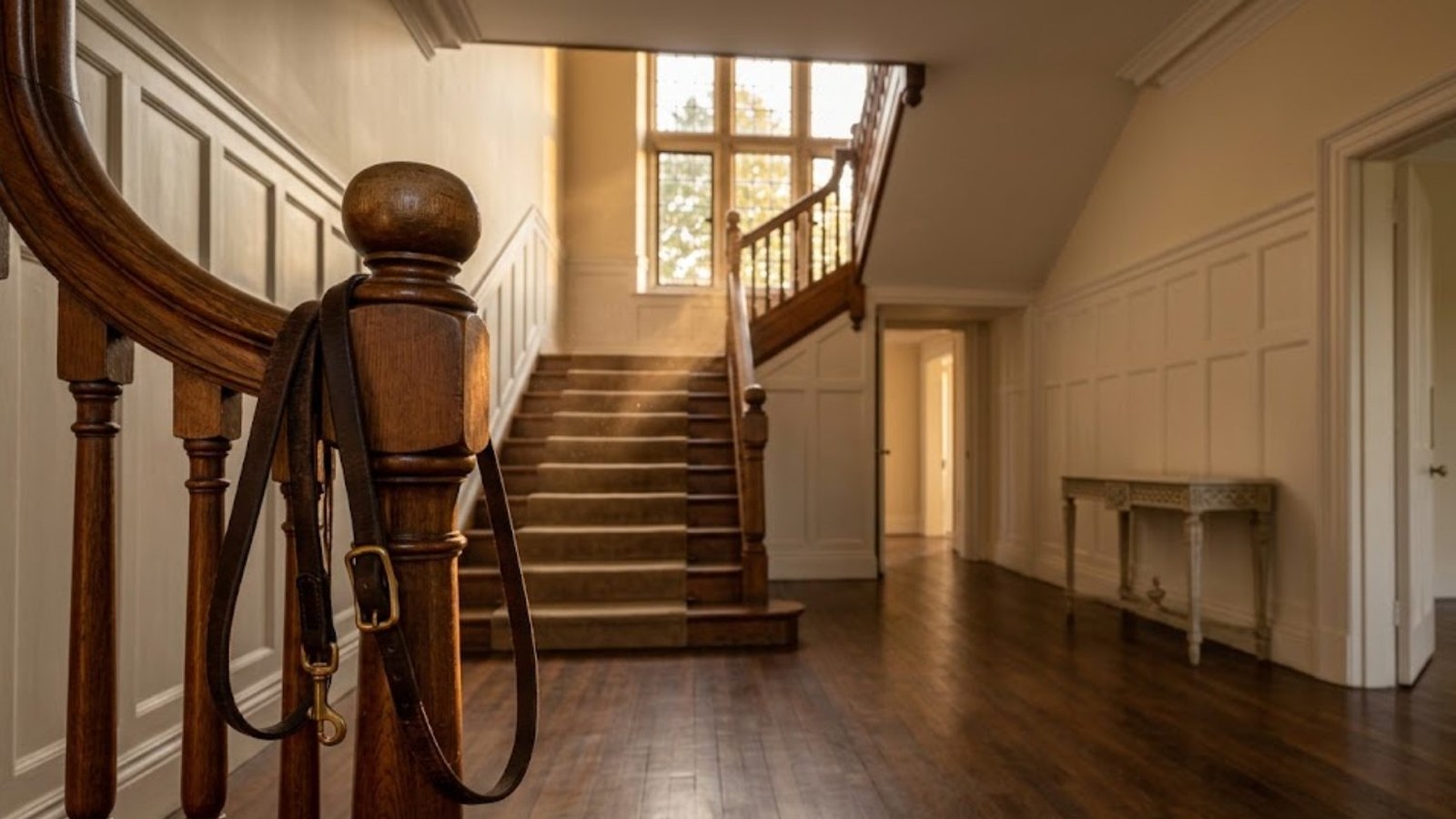 An empty house's front hall in warm afternoon light, a dog leash hanging from the newel post of a wooden staircase, bare floors and one console table.