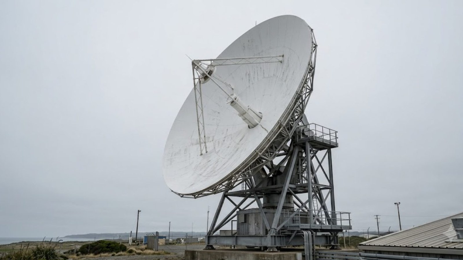 A large white dish antenna on a steel lattice mount at a coastal ground station, tilted toward a flat overcast sky above scrub and low metal buildings.