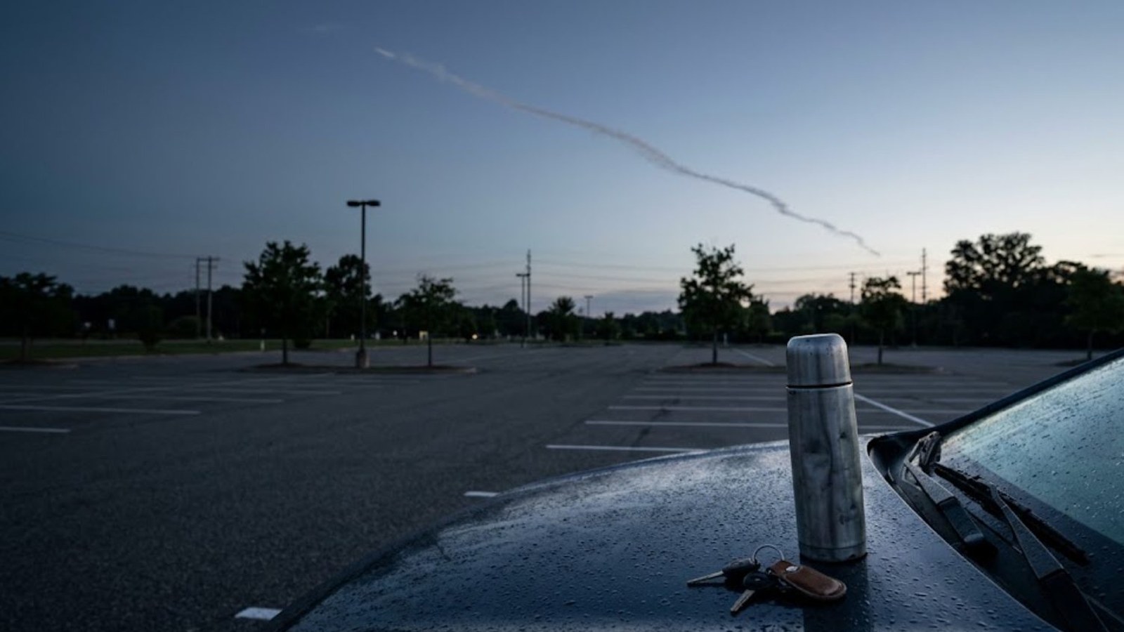 A steel thermos and car keys rest on the rain-beaded hood of a car in an empty parking lot at dusk, a thin vapor trail crossing the darkening sky.