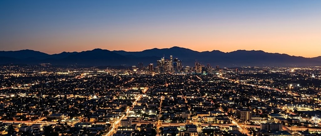 Los Angeles spreads out in lights at dusk below a dark mountain ridge, the downtown towers glowing under an orange and deep blue sky.