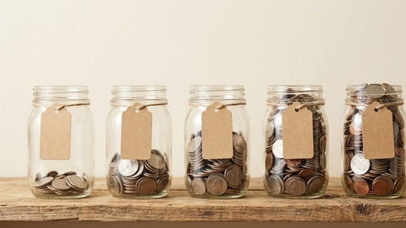 Five glass jars with blank paper tags sit in a row on a wooden shelf, each holding a larger pile of coins than the last.