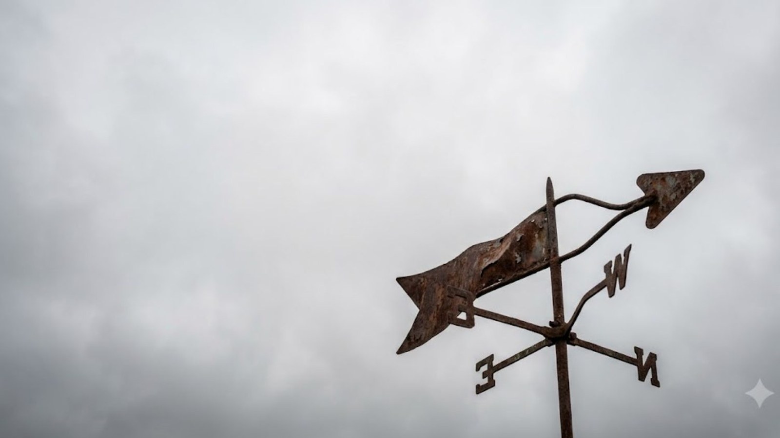 A rusted weathervane with an arrow and compass letters points into a low gray sky of gathering storm clouds.