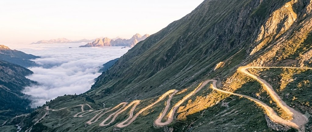 A winding mountain road climbs in tight switchbacks across a green slope at sunrise, a sea of clouds filling the valley beyond.