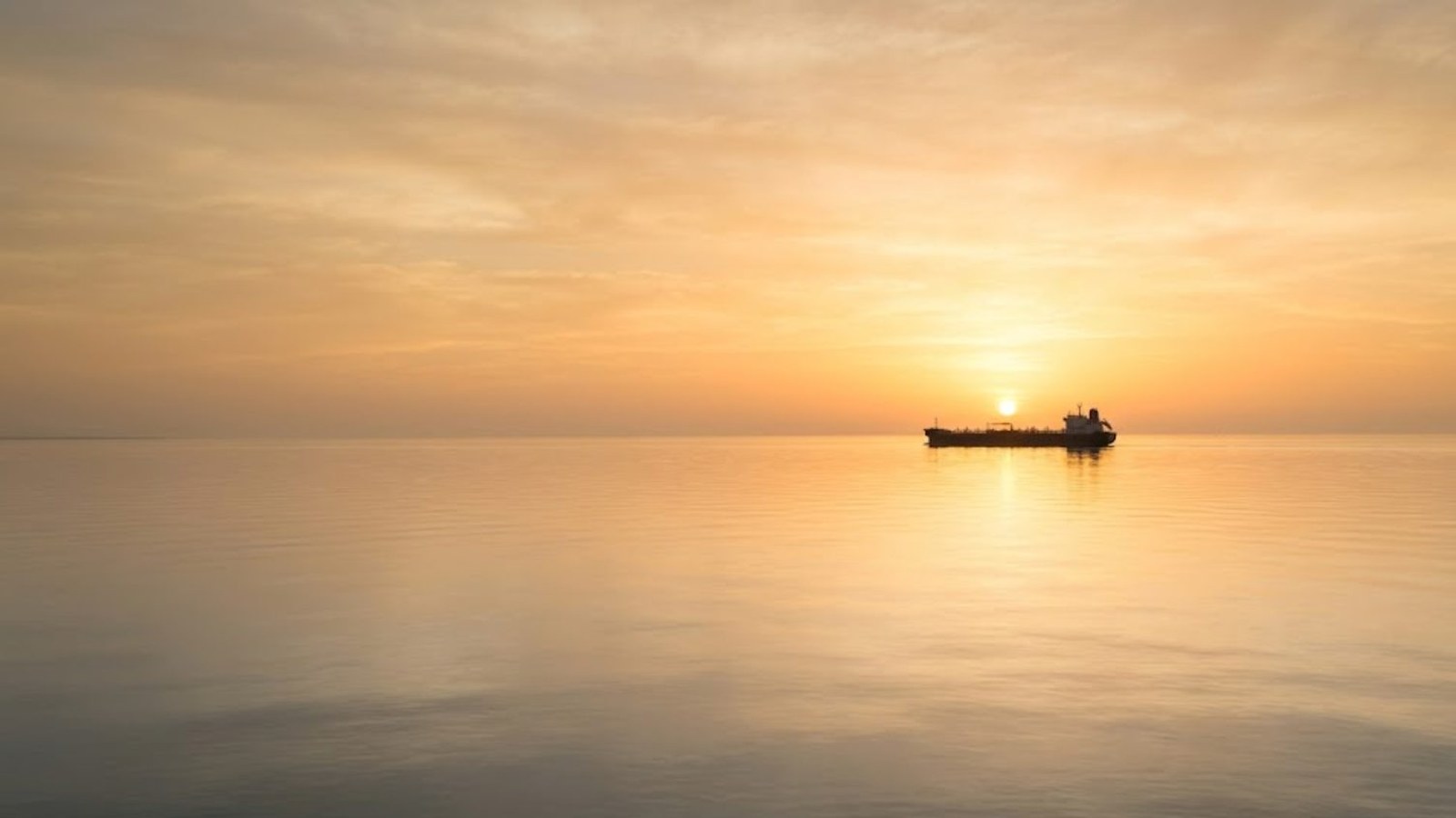 A crude tanker sits in silhouette on a flat, calm sea as the sun rises through amber haze on the horizon.
