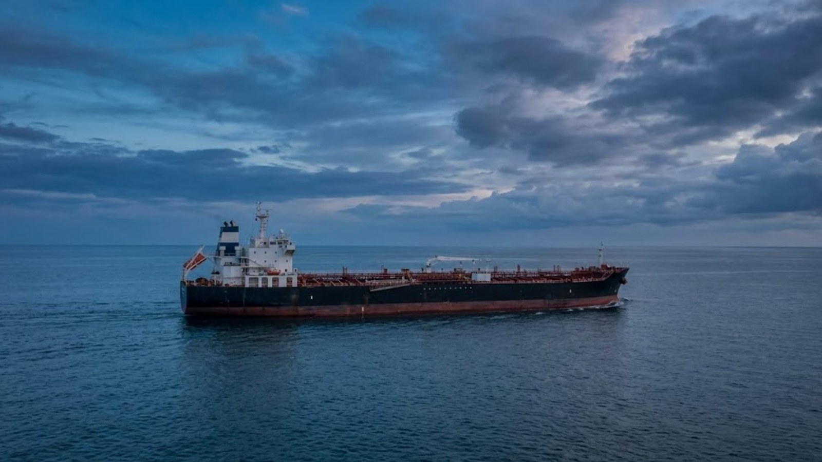 An oil tanker with a dark red hull crosses calm open water under heavy blue clouds at dusk, seen from above and off its bow.