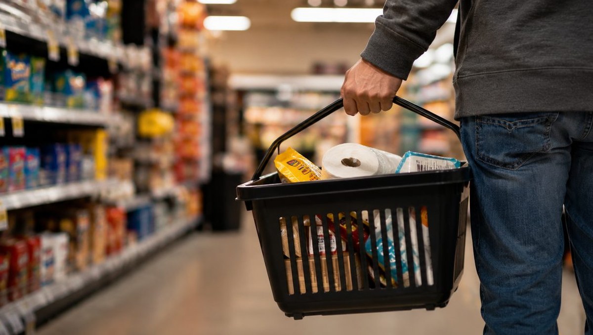 A shopper in jeans carries a black plastic basket of paper towels and groceries down a brightly lit store aisle, shelves blurred on either side.