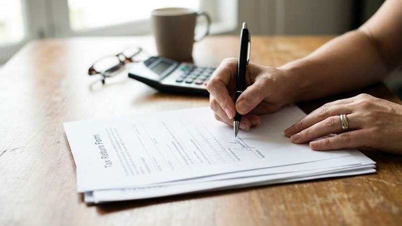 A hand signs a stack of papers on a worn wooden table beside a calculator, reading glasses and a coffee mug in soft window light.
