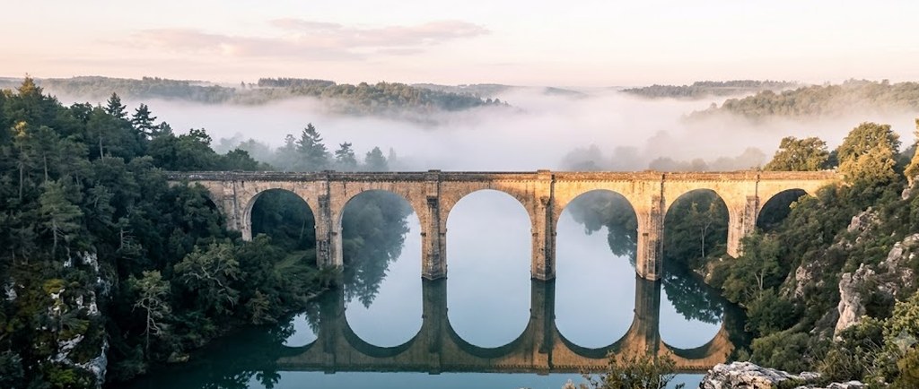 A many-arched stone bridge spans a still river in a wooded valley at dawn, its arches mirrored in the water and mist drifting over the hills.