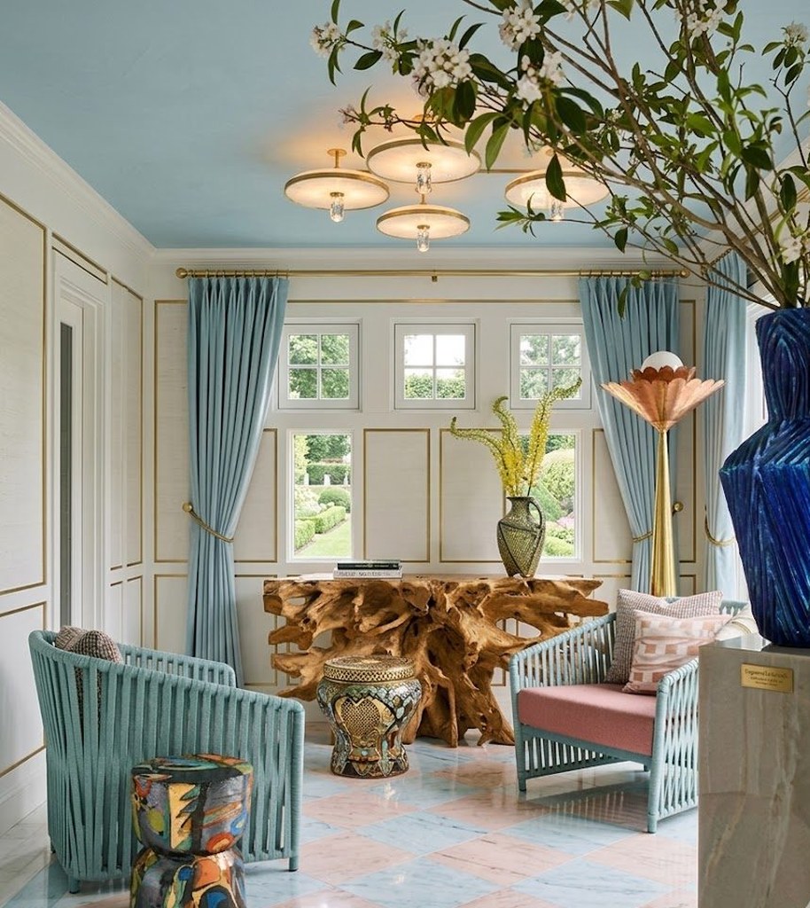 A bright formal sitting room with pale blue curtains, sculpted armchairs, a carved wooden console and a tall cobalt vase holding a leafy branch.