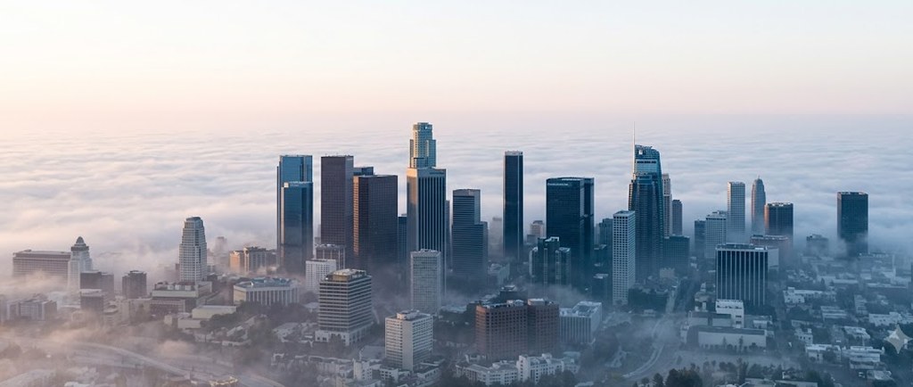 Downtown Los Angeles towers rise out of a thick bank of low morning fog seen from above, the surrounding streets fading into pale haze at dawn.