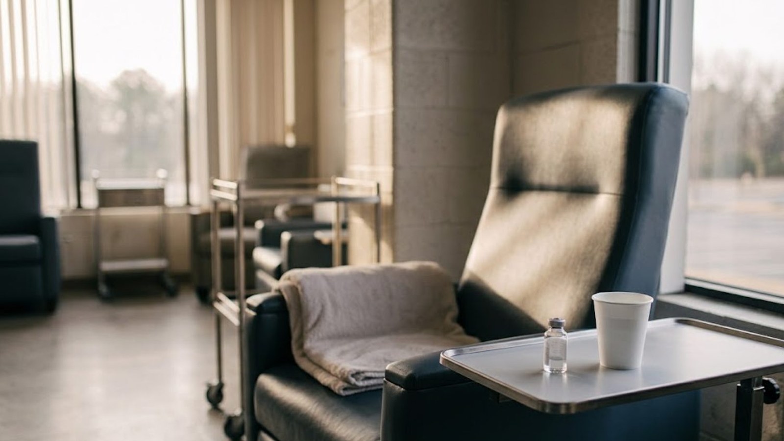 An empty dark leather treatment recliner sits by a tall window in a quiet clinic, a folded blanket on the seat, a paper cup and vial on the tray.