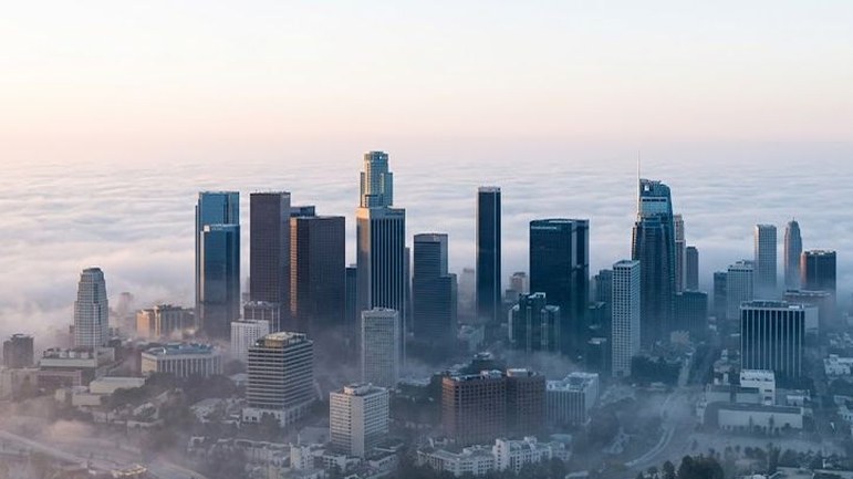 A cluster of downtown towers rises out of a thick layer of morning fog, seen from the air in soft pale light, the city below half hidden.
