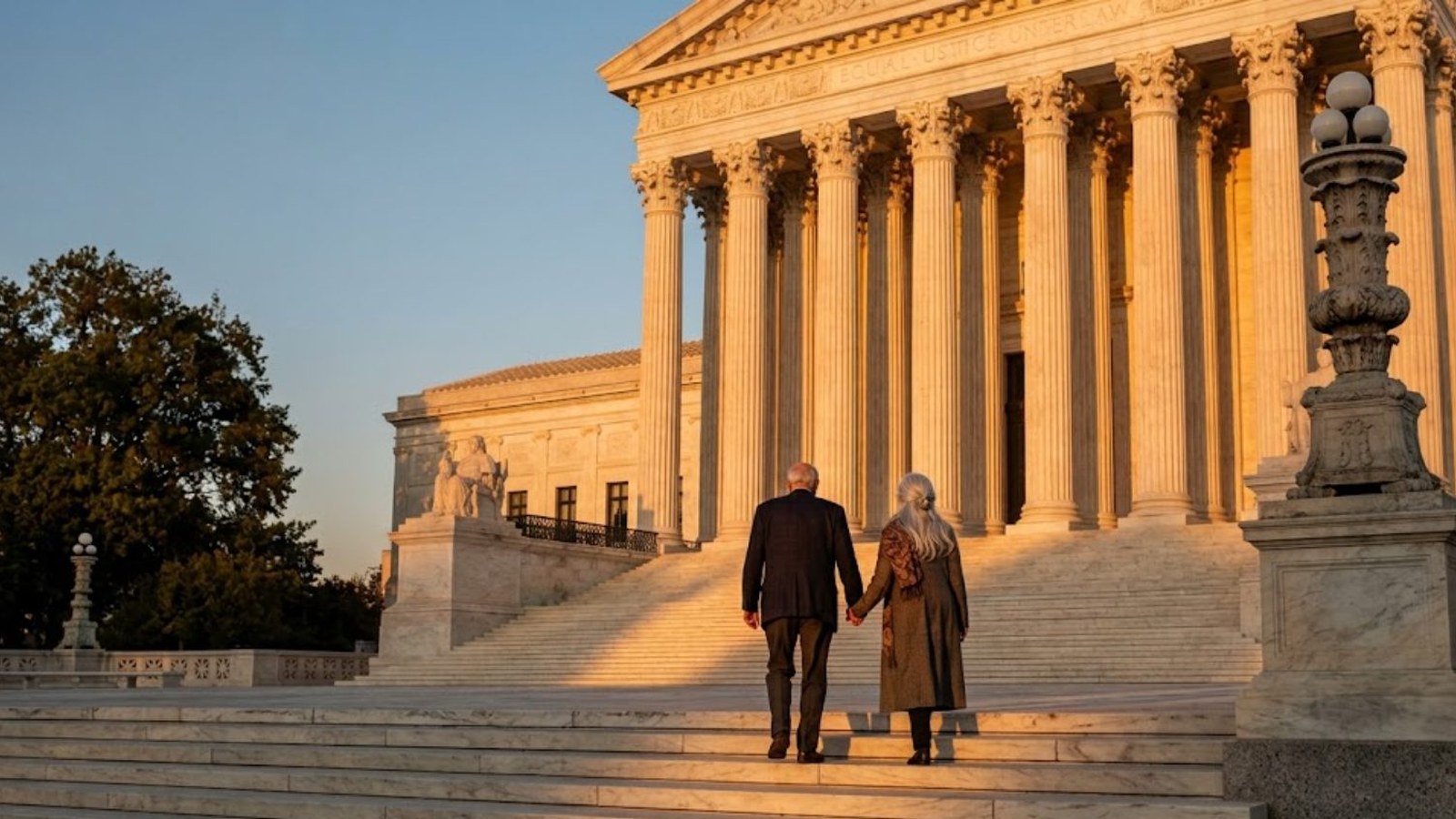 An older couple seen from behind, holding hands as they climb the wide marble steps of a columned government building in warm early-evening light.