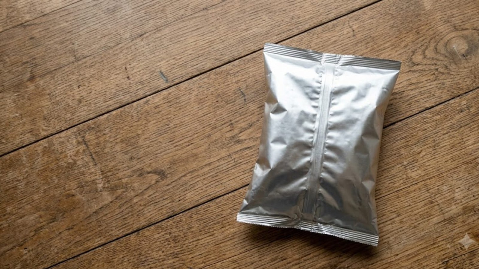 A single unbranded silver foil snack bag lying on a bare wooden counter, shot from directly overhead in soft even light.