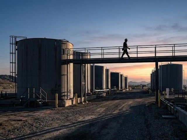 A worker in a hard hat crosses a steel walkway between tall fuel storage tanks at sunrise, a gravel yard in shadow below and hills beyond.