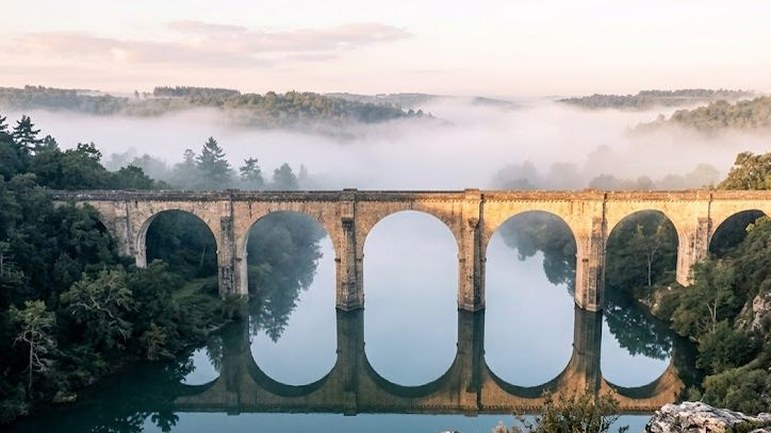 An old stone viaduct of many arches crosses a still river in a wooded valley, morning fog lying over the hills and the arches mirrored in the water.