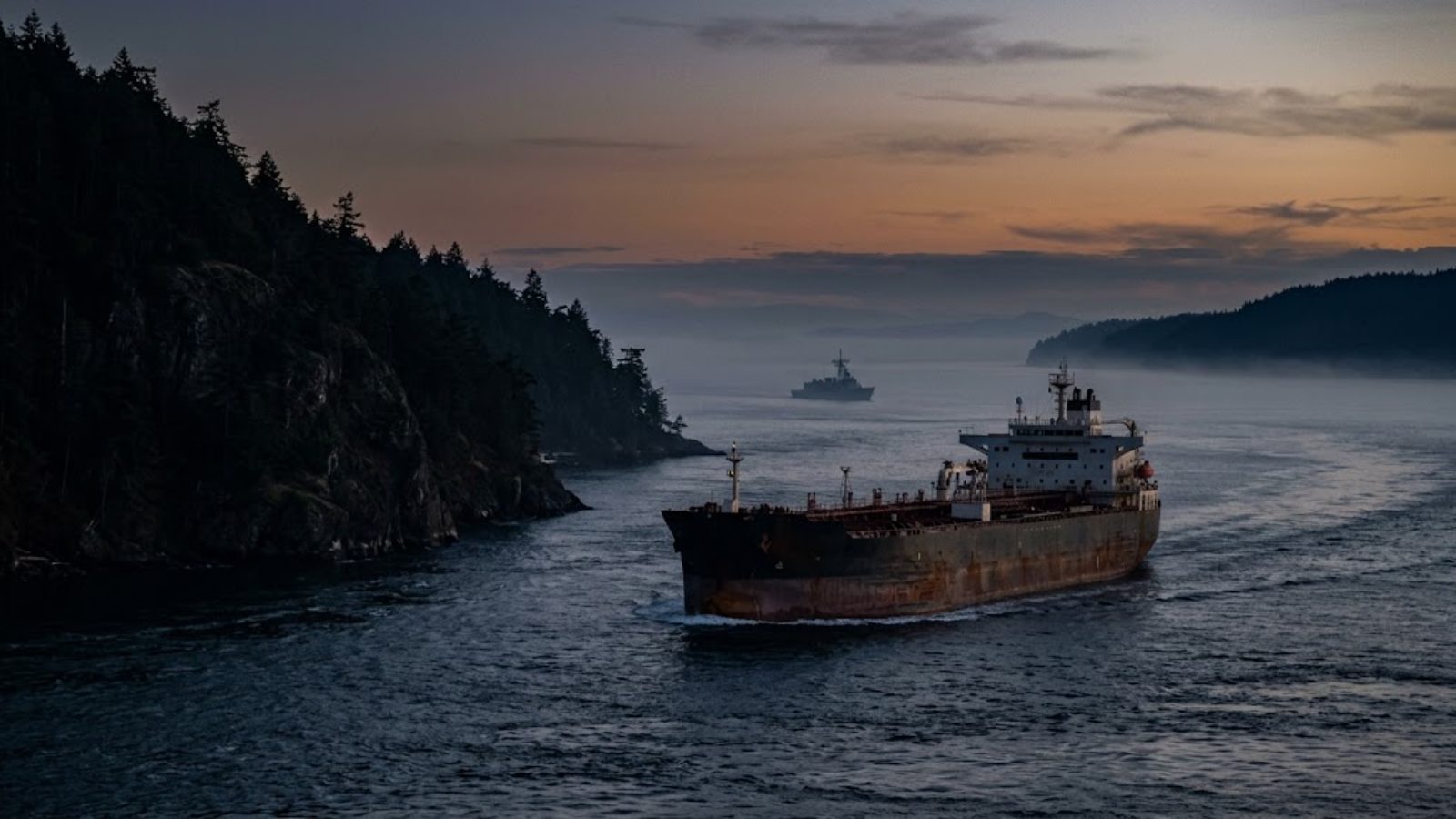 A worker crossing a catwalk above rows of crude storage tanks at dusk — the toll on oil that already moves.