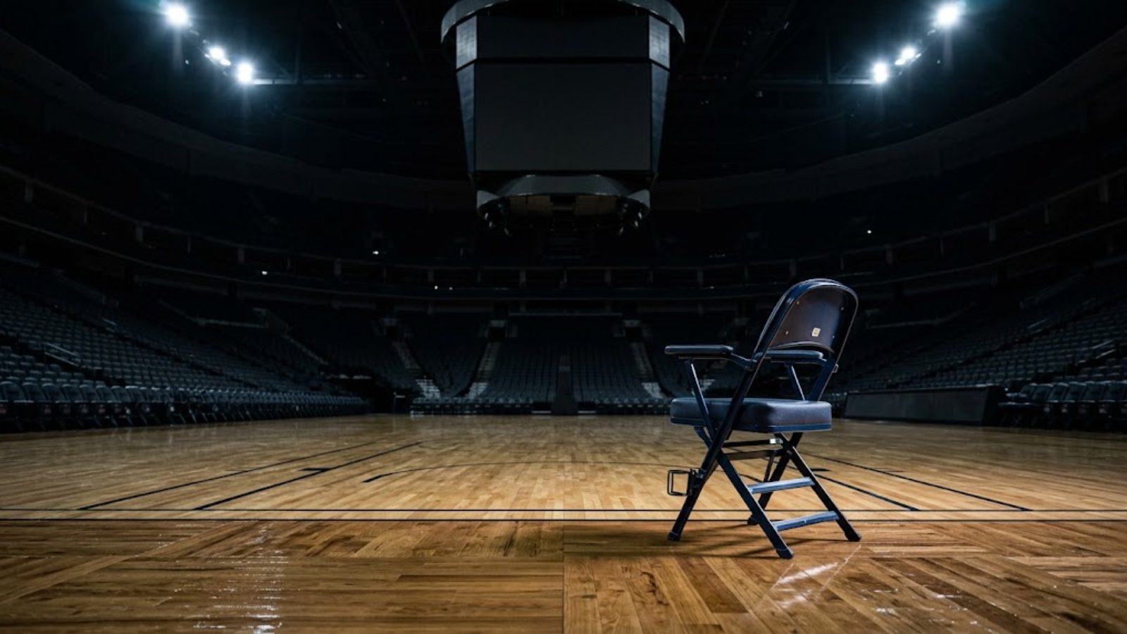 An empty courtside seat under bright arena lights beside a polished hardwood floor, the stands dark and unlit behind it.