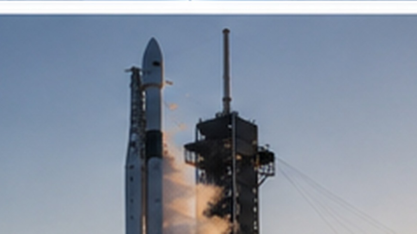 A rocket standing on its launch pad beside the service tower, seen against an empty pale sky