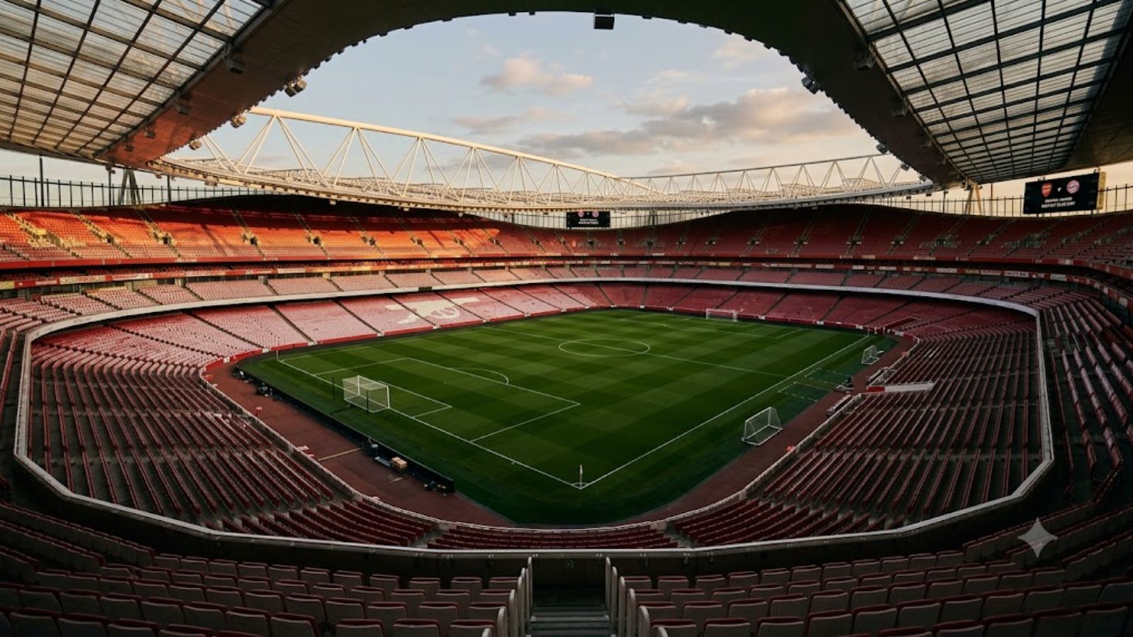 An empty stadium bowl at dusk, red seats sweeping around a floodlit green field, no one on the pitch.
