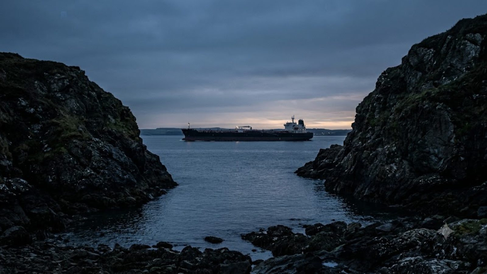 A laden tanker anchored in a rocky cove at dusk, hills dark on either side.