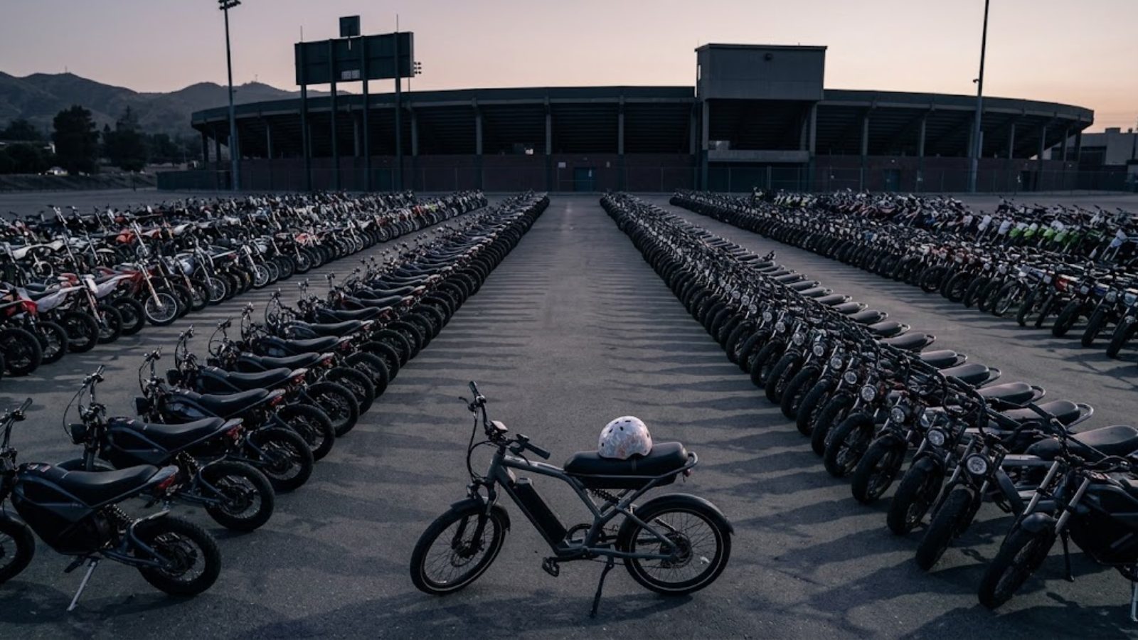 Rows of e-bikes and small motorcycles lined up in a stadium parking lot at dusk.