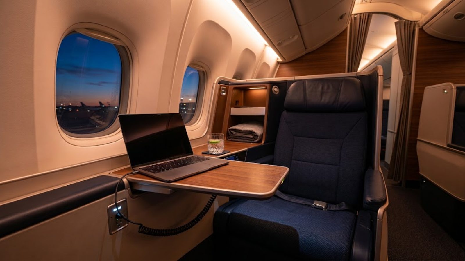 A wide first-class seat with a laptop open on the tray table, sunrise outside the window.