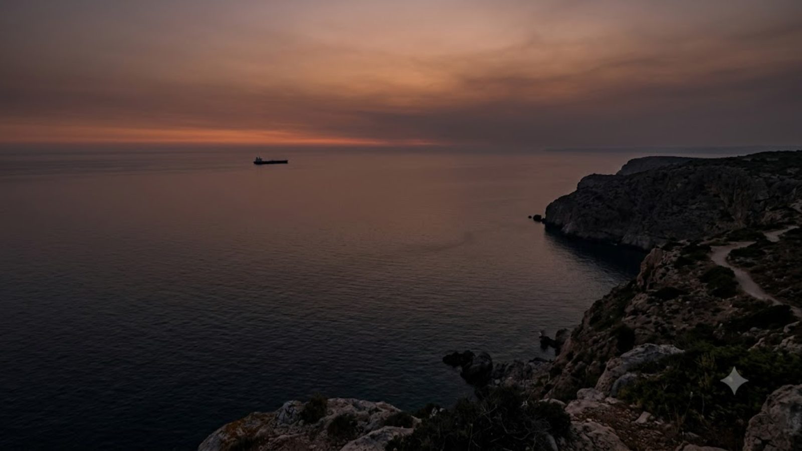 A lone ship on the horizon off a rocky coastline at dusk.